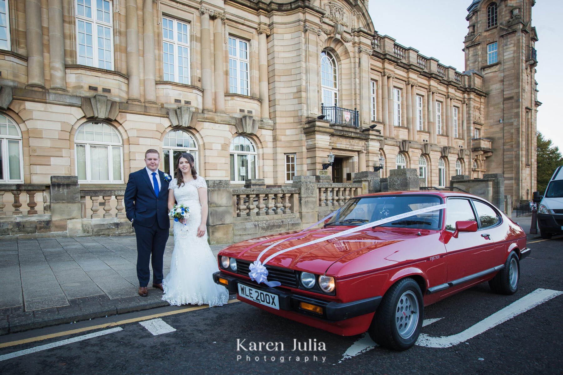 bride and groom pose for a portrait with their wedding car, a red Ford Capri