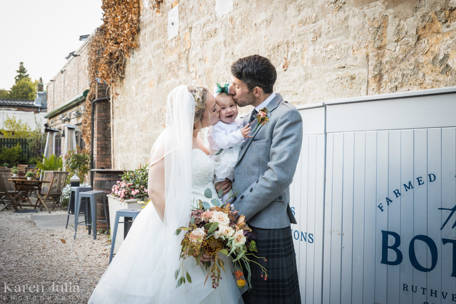 bride and groom portrait kissing the chubby cheeks of their baby at the entrance to the Bothy